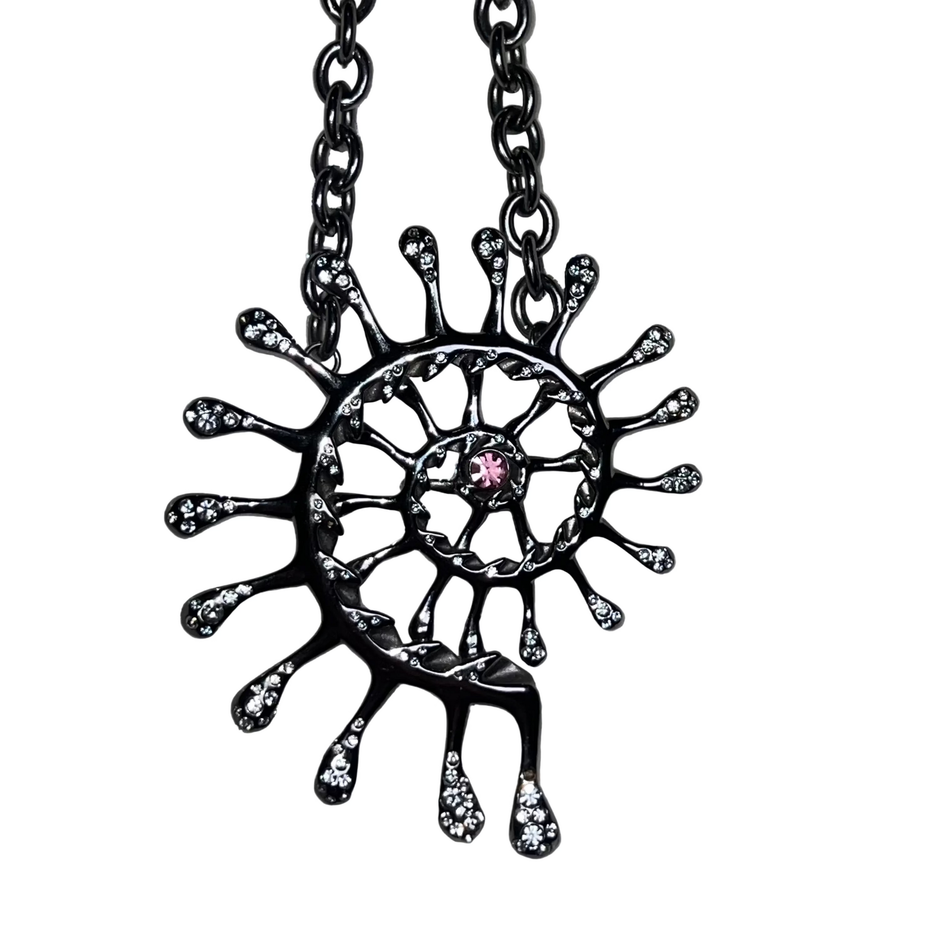 A black sun pendant necklace with small clear gemstones and a central pink gemstone, hanging from a black chain on a shiny silver fabric background.