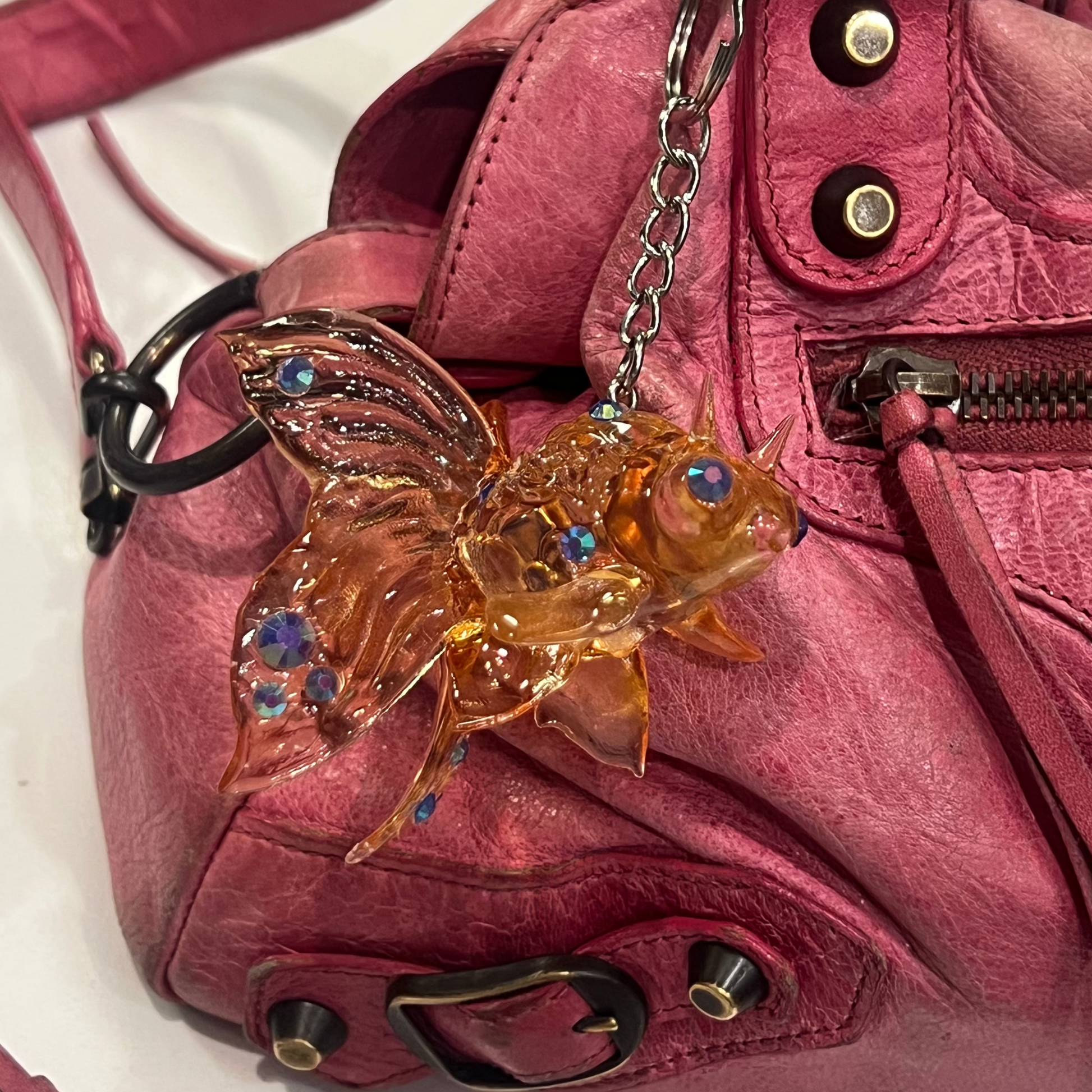 A close-up of a pink leather handbag featuring a decorative amber-colored fish charm with blue rhinestone accents hanging from a metal chain.