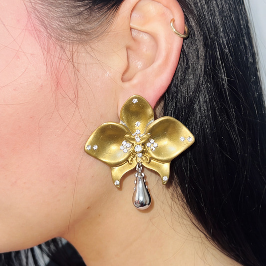 A close-up side view of a person's ear wearing a large gold orchid-shaped earring with small clear stones and a dangling silver teardrop charm. A small gold hoop earring is also visible on the ear.