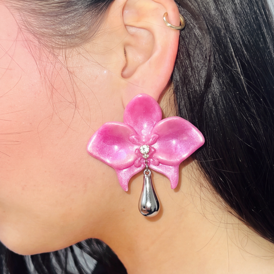 A close-up of a person's ear wearing a large pink orchid flower earring with a dangling silver teardrop charm and a small clear rhinestone at the center.