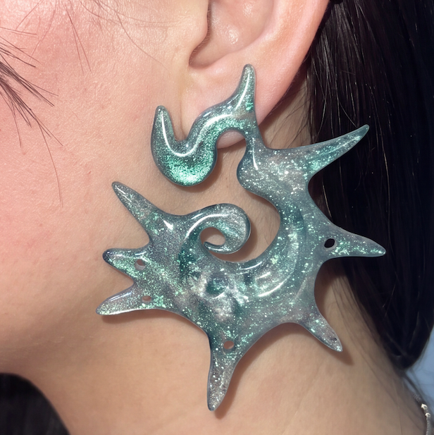 A close-up of a person's ear wearing a large, organic-shaped earring made of iridescent, glittery mint green and silver material.