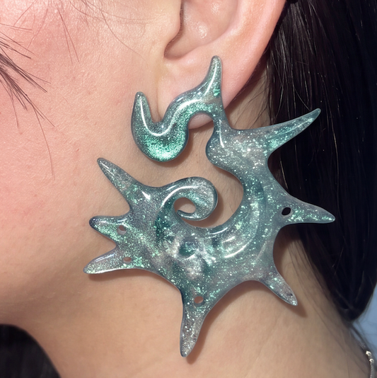 A close-up of a person's ear wearing a large, organic-shaped earring made of iridescent, glittery mint green and silver material.