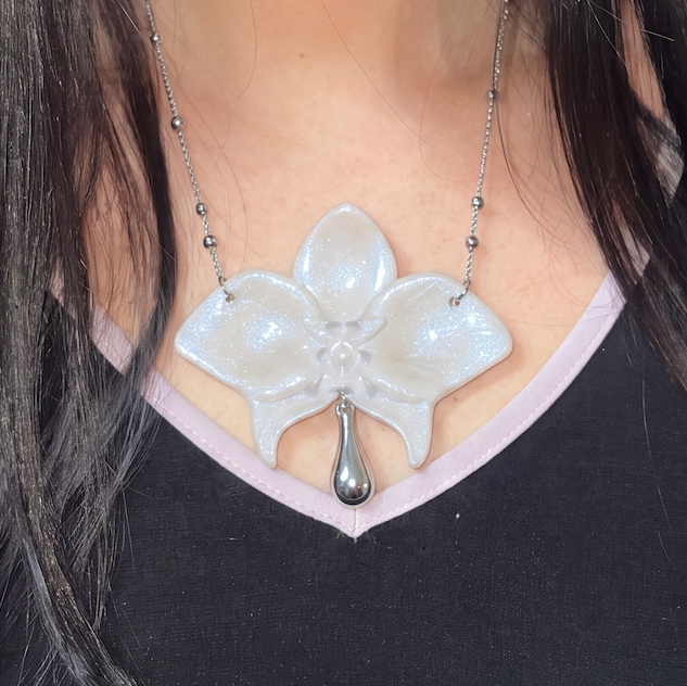 A close-up of a person wearing a silver orchid flower pendant necklace with a dangling teardrop charm, styled over a black and light pink V-neck top.
