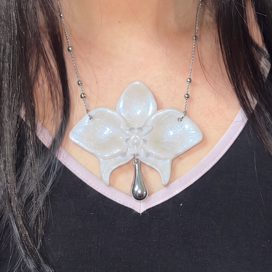 A close-up of a person wearing a silver orchid flower pendant necklace with a dangling teardrop charm, styled over a black and light pink V-neck top.